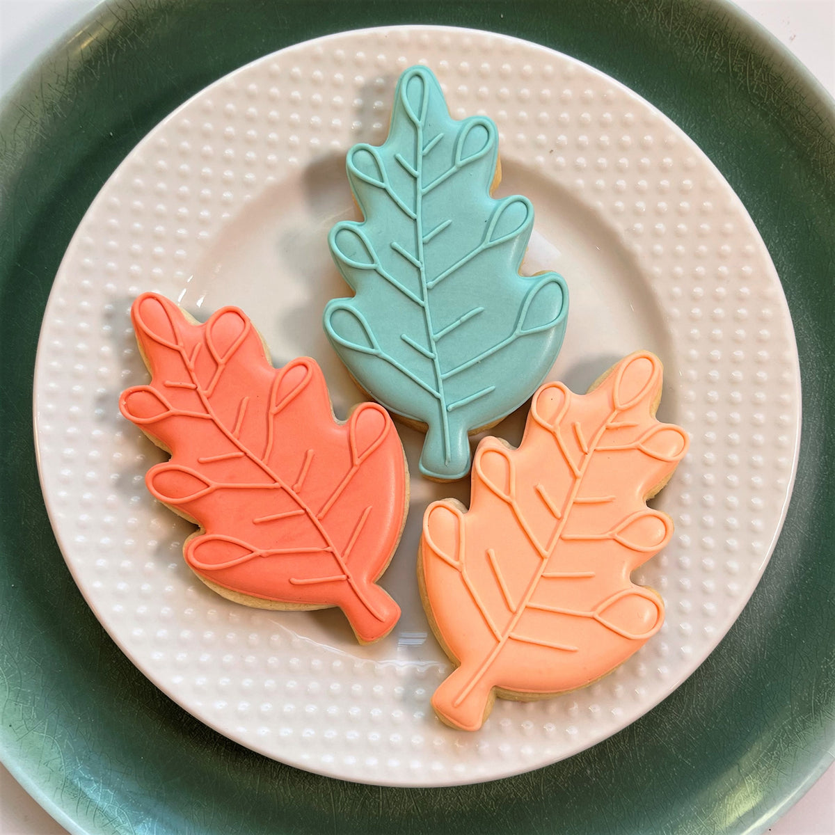 Three coral peach and teal autumn leaf sugar cookies decorated with detailed veins made using leaf doodle cookie cutter