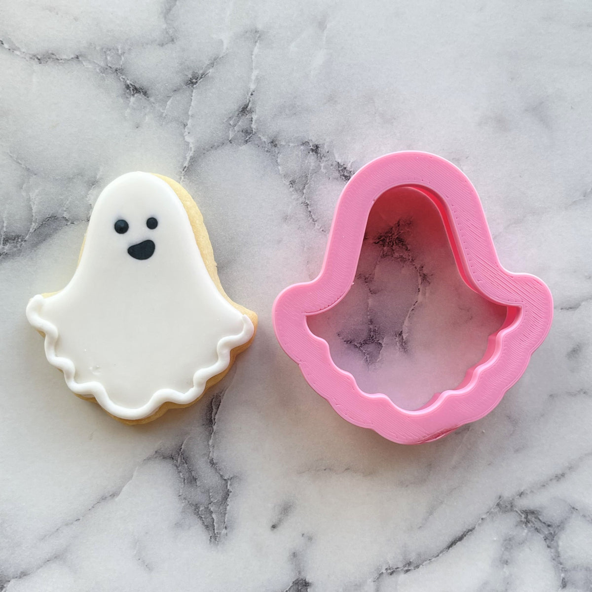 Mini classic ghost cookie cutter in pink plastic next to a white frosted ghost cookie with black icing details