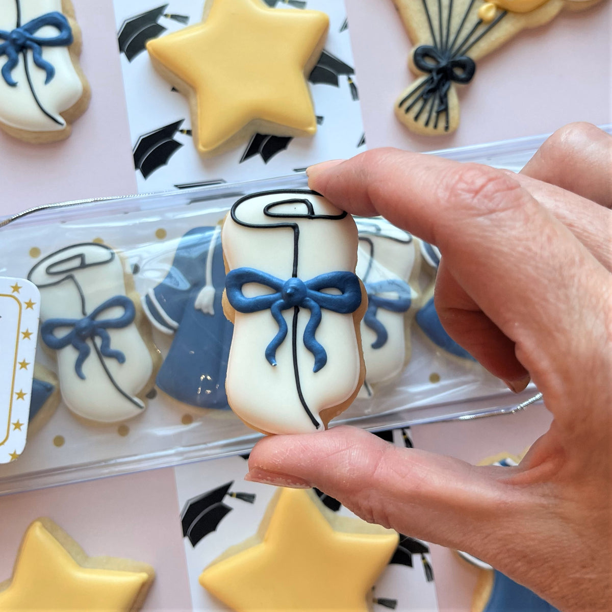 A hand holds a white frosted graduation cap cookie with a blue icing bow made using the mini grad cookie cutter bundle