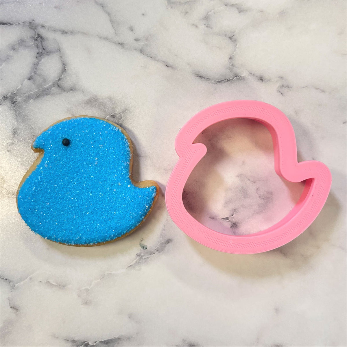 A vibrant blue glittery bird shaped cookie next to a pink plastic mini easter chick cookie cutter