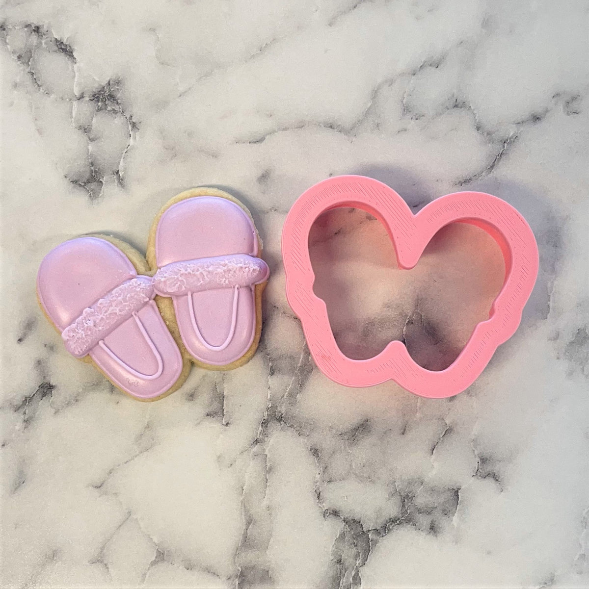Pink heart-shaped mini slippers cookie cutter next to two pink slipper cookies with lace detailing on a white background