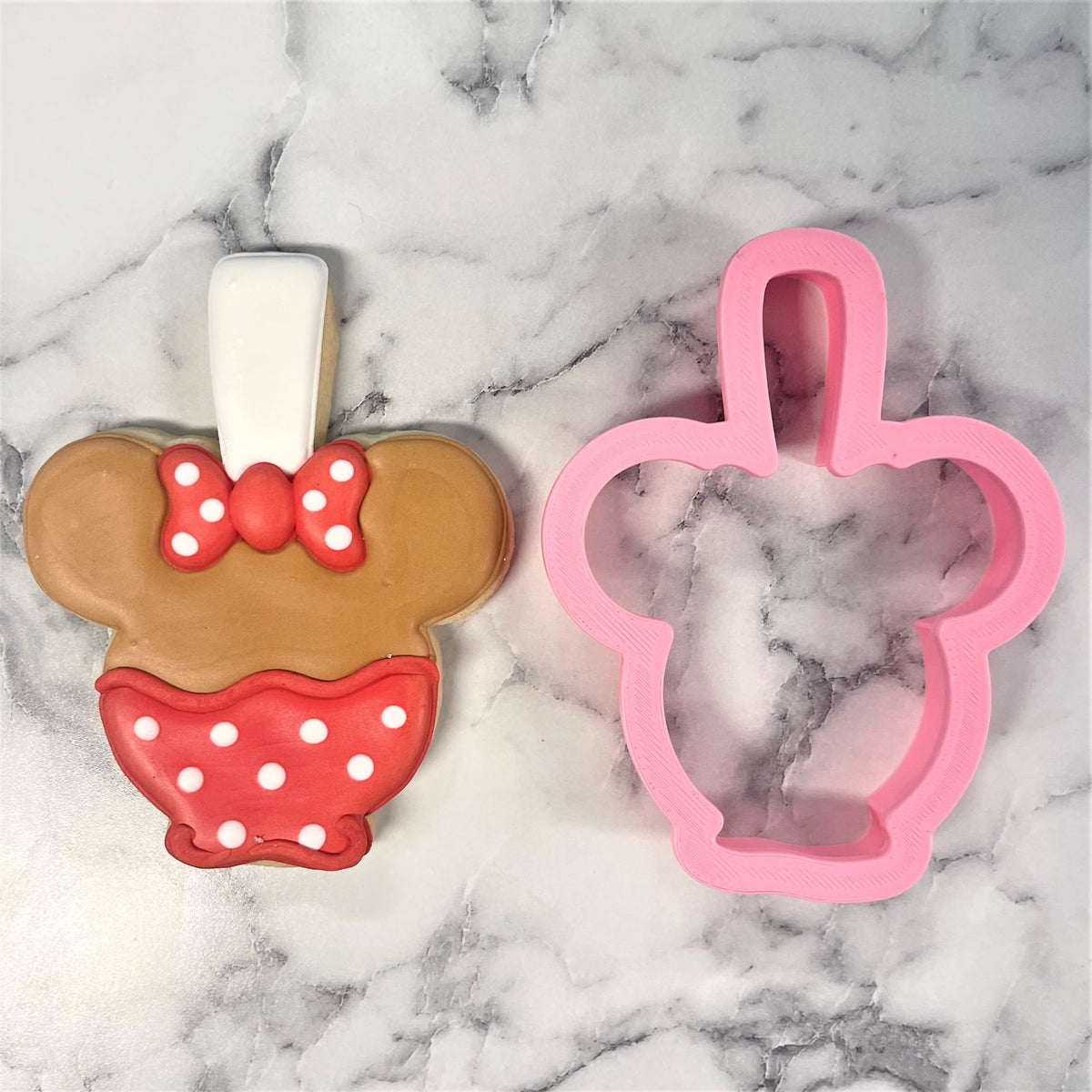 Pink plastic minnie mouse-shaped mouse candy apple cookie cutter next to a decorated matching cookie