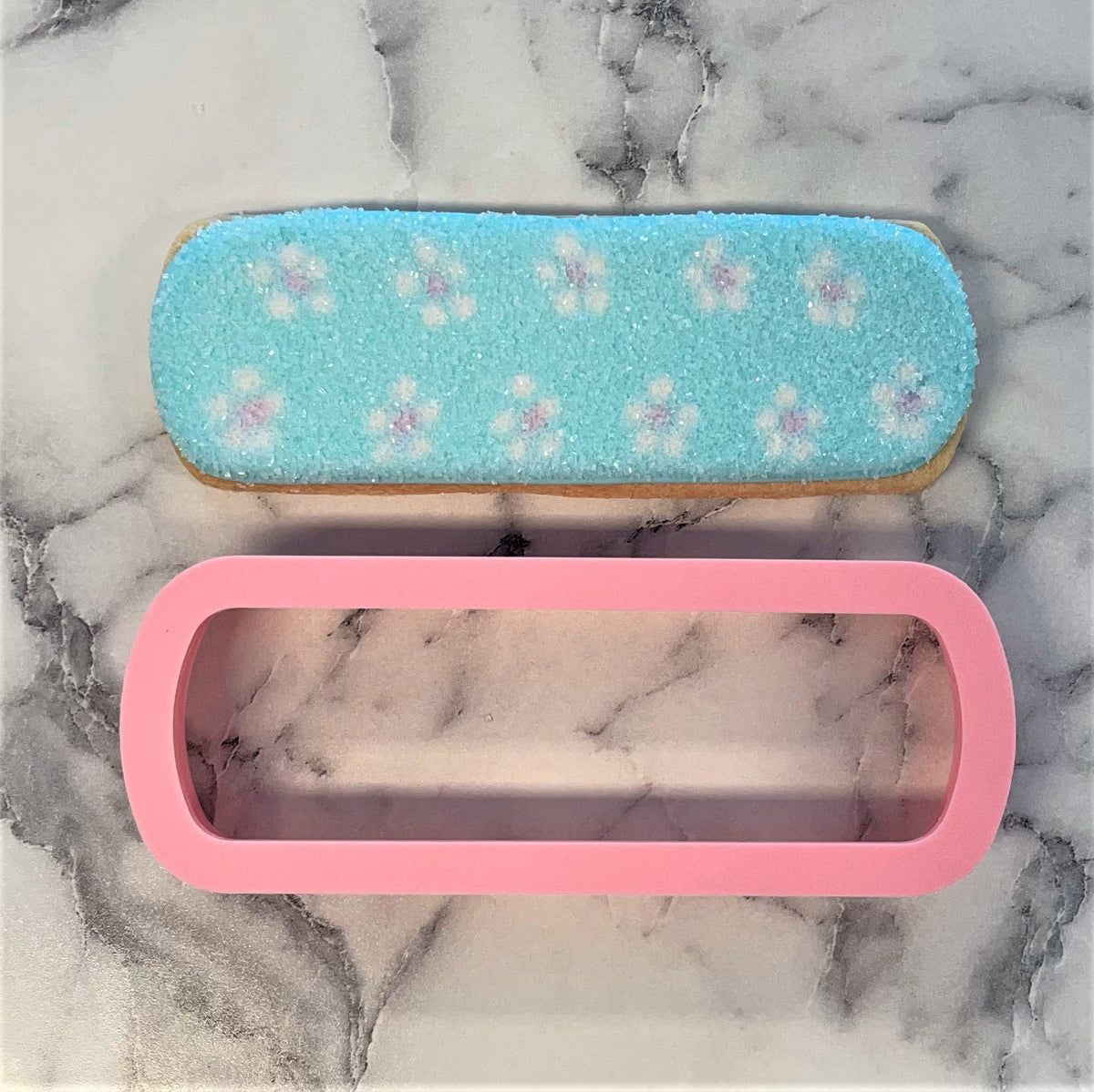A pink silicone nail file cookie cutter with a rounded rectangular shape beside a light blue frosted floral cookie