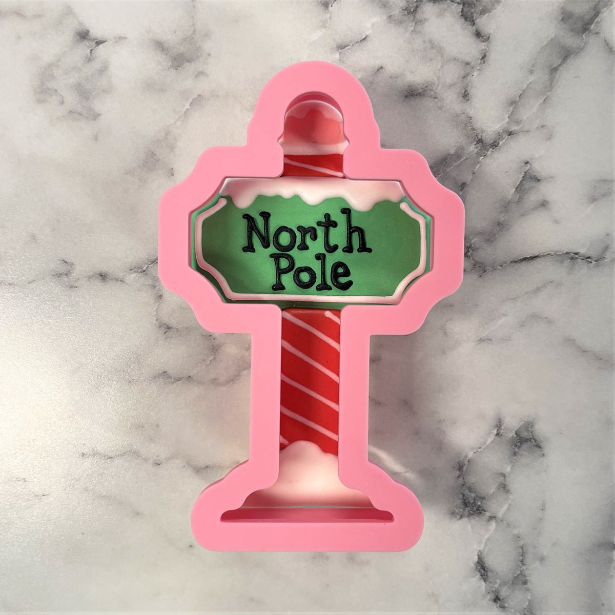 Pink silicone north pole sign cookie cutter shaped like a candy cane pole with a green north pole sign