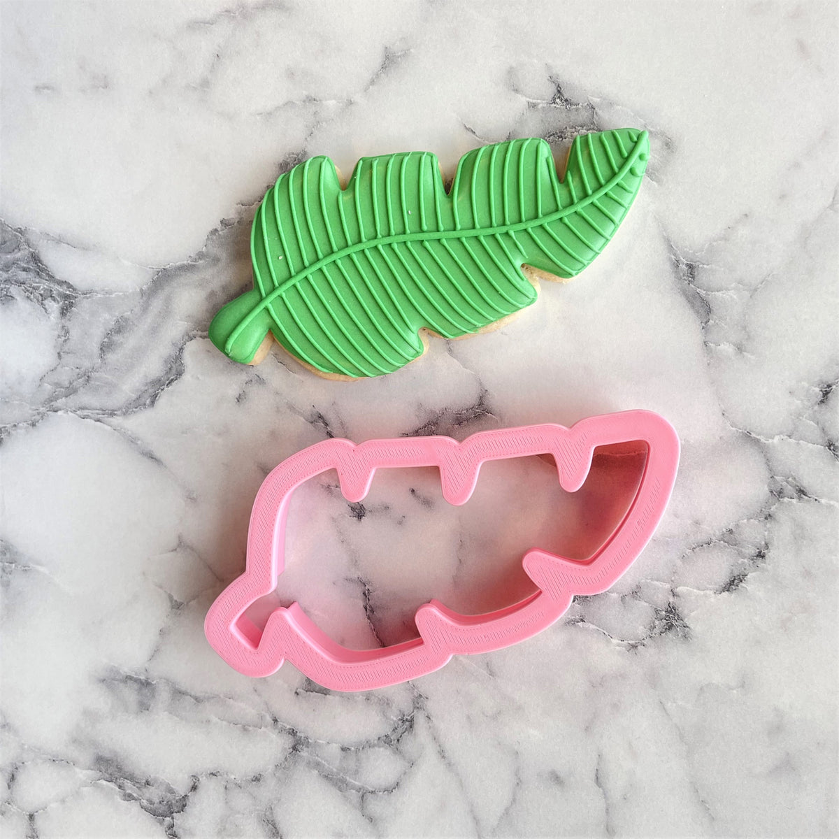 Green leaf-shaped palm branch cookie with detailed vein patterns and pink plastic cookie cutter beside it
