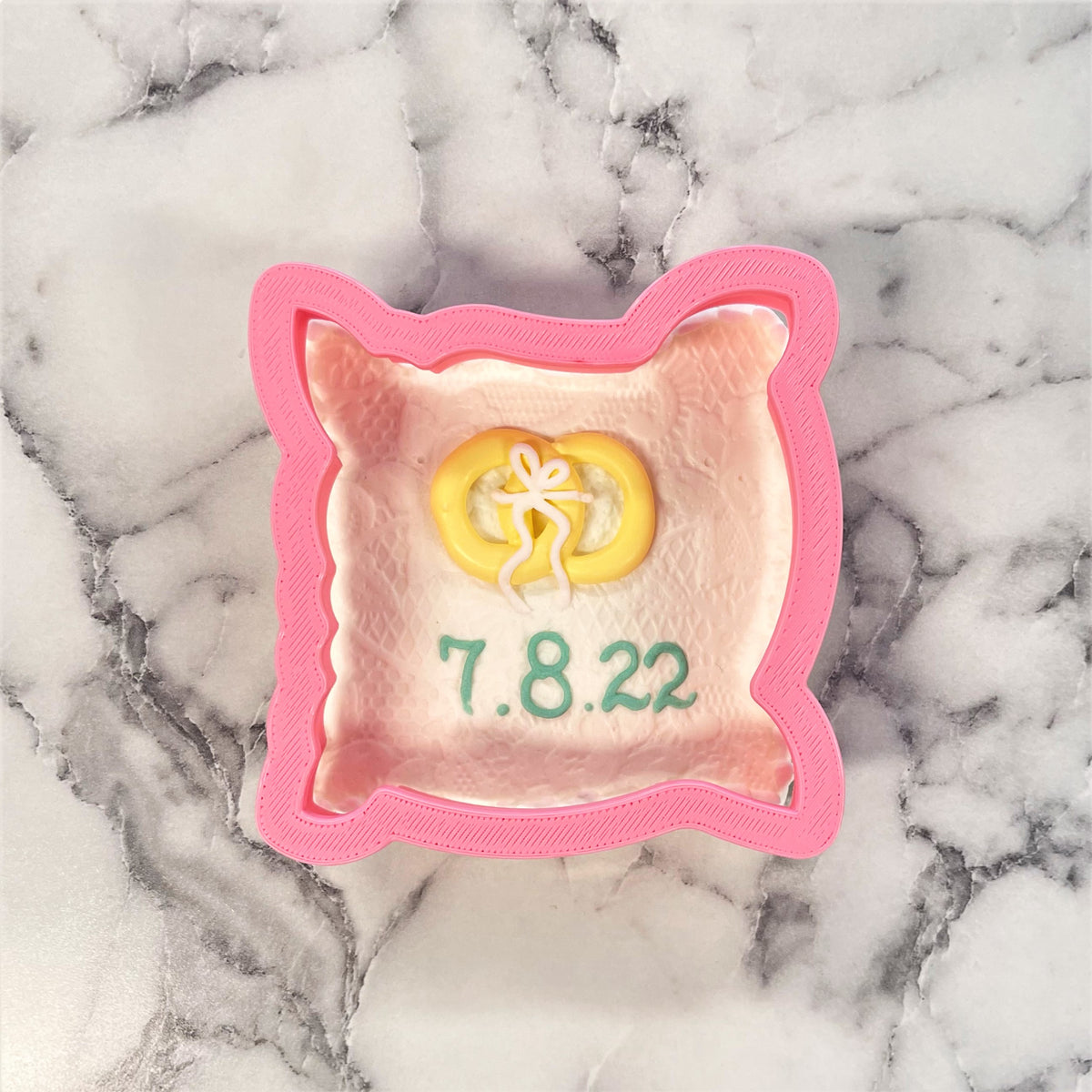 Pink pillow cookie cutter shaped like cat ears with pink interior white icing and green text reading 7.8.22