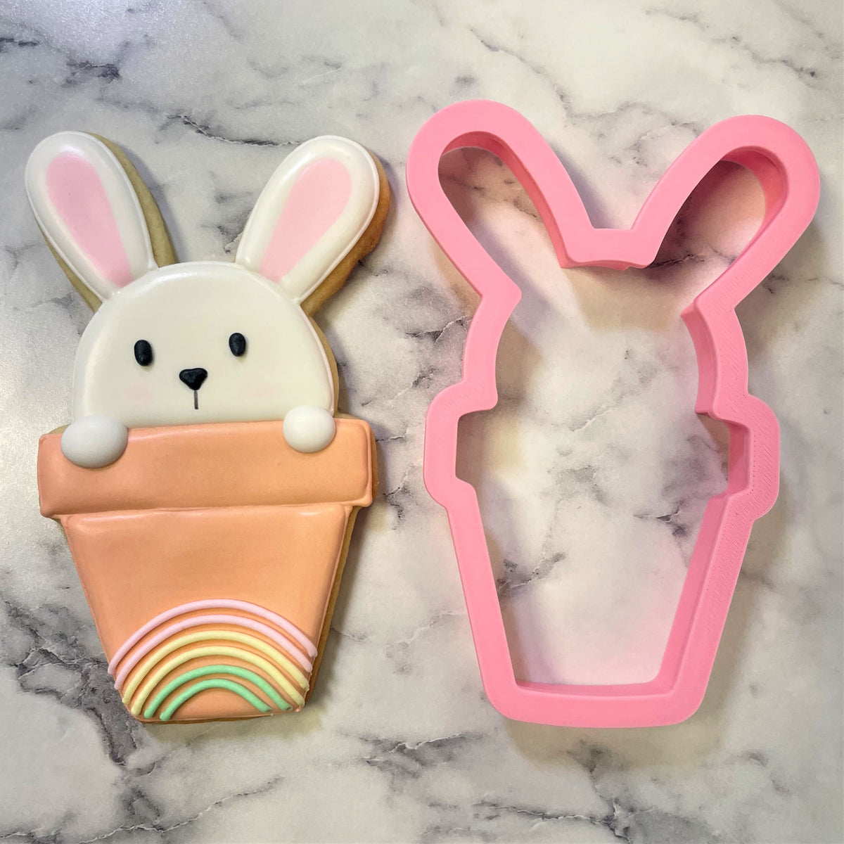 Pink bunny-shaped cookie cutter next to a decorated sugar cookie with rainbow icing in a pot design