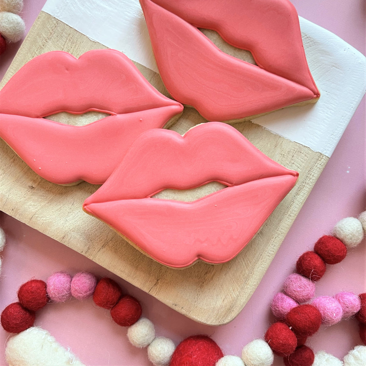 Three pink-frosted sugar cookies shaped like lips arranged on a wooden board made using puckered lips cookie cutter