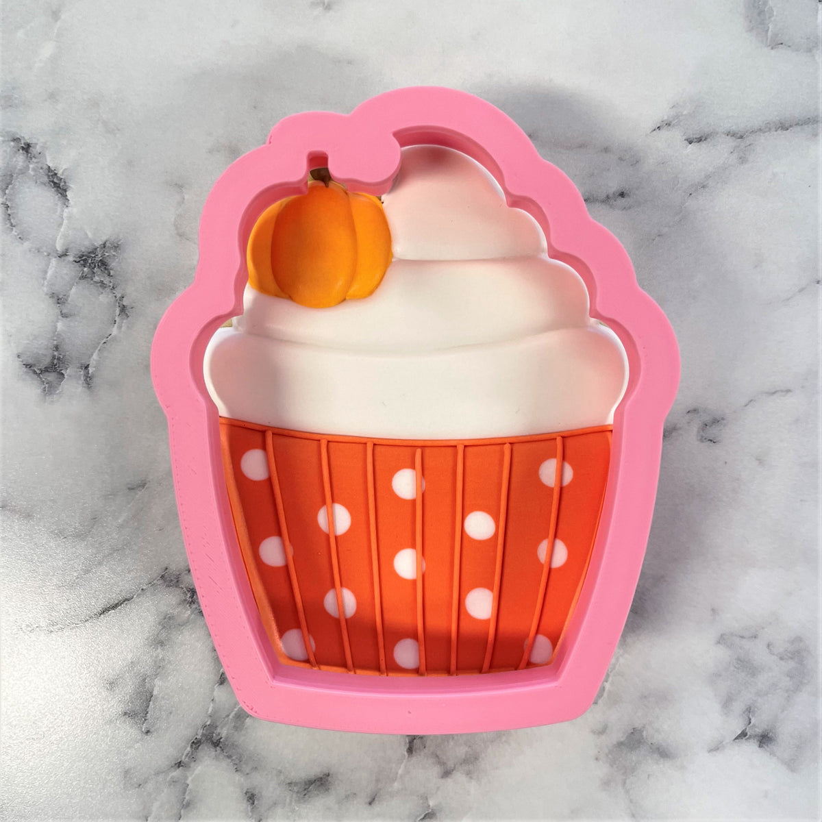 Pink silicone pumpkin cupcake cookie cutter with orange polka-dot liner and white frosting detail topped with small pumpkin