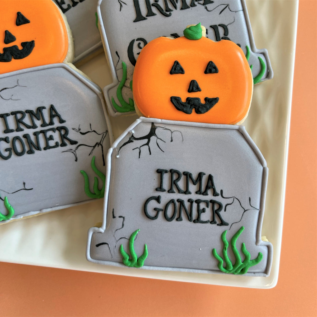 Orange pumpkin-shaped cookie with black icing face and green stem on gray tombstone cookie in haunted house decorating class recording