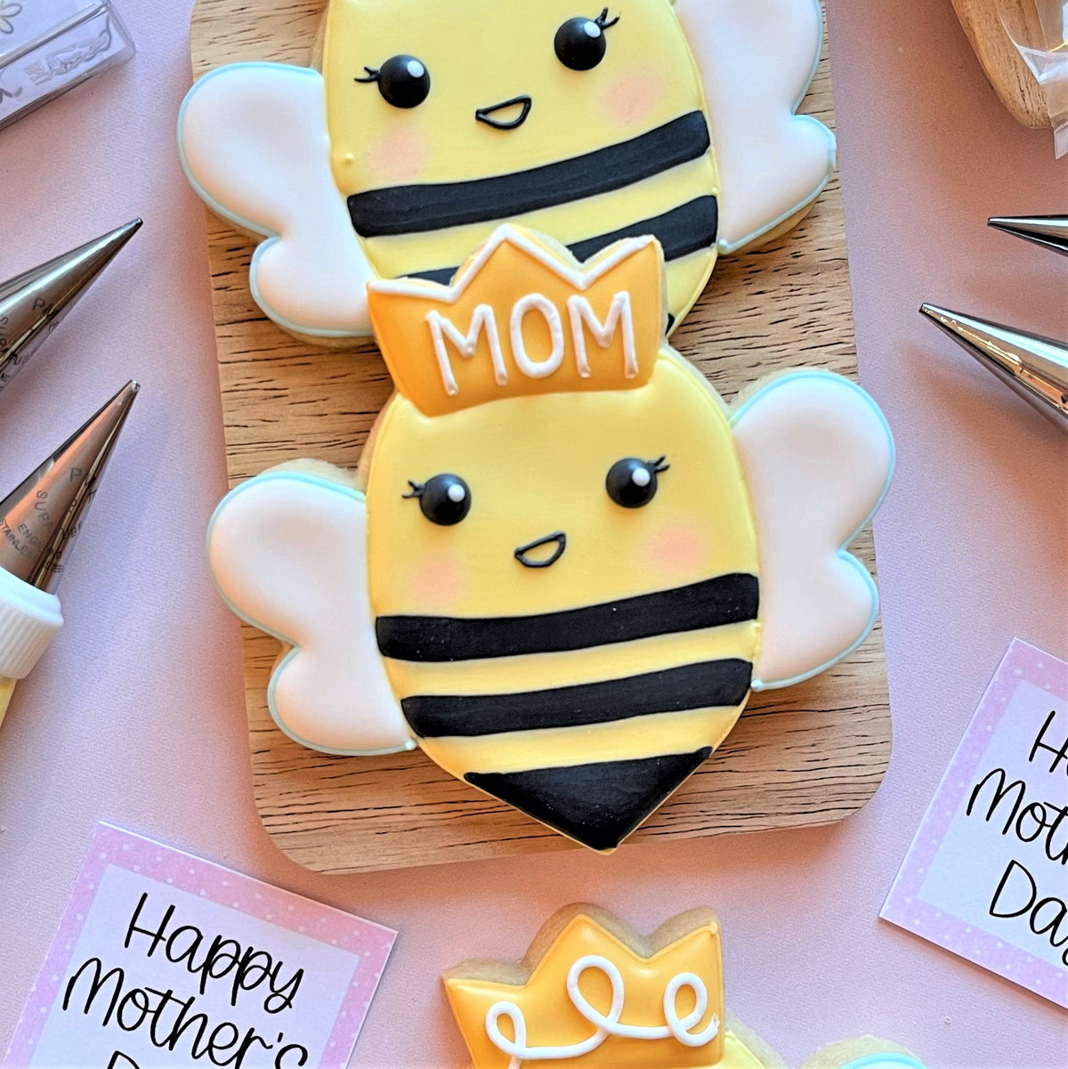 Yellow and black striped bee-shaped cookies with white wings and smiling faces wearing golden mom crowns made using queen bee cookie cutter