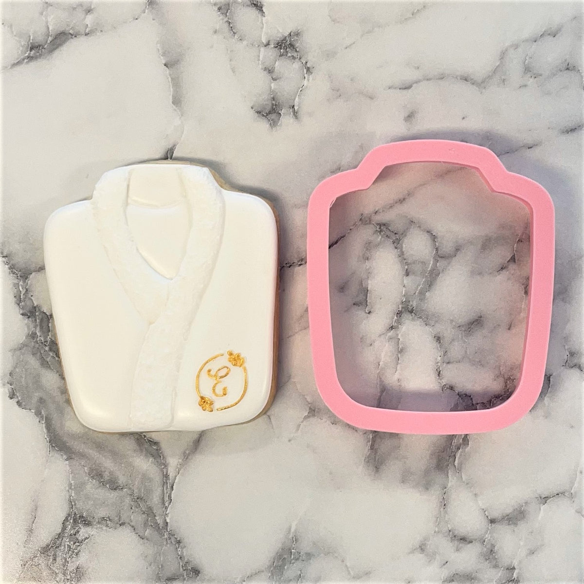 White bathrobe-shaped cookie with gold monogram detail next to pink silicone robe cookie cutter for baking use