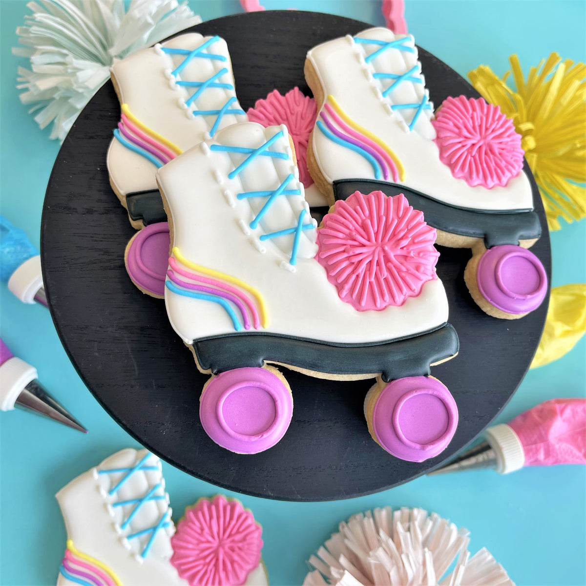 White frosted roller skate cookies with blue lace details rainbow stripes pink pom pom accents and purple wheels for totally 80s cookie decorating class recording