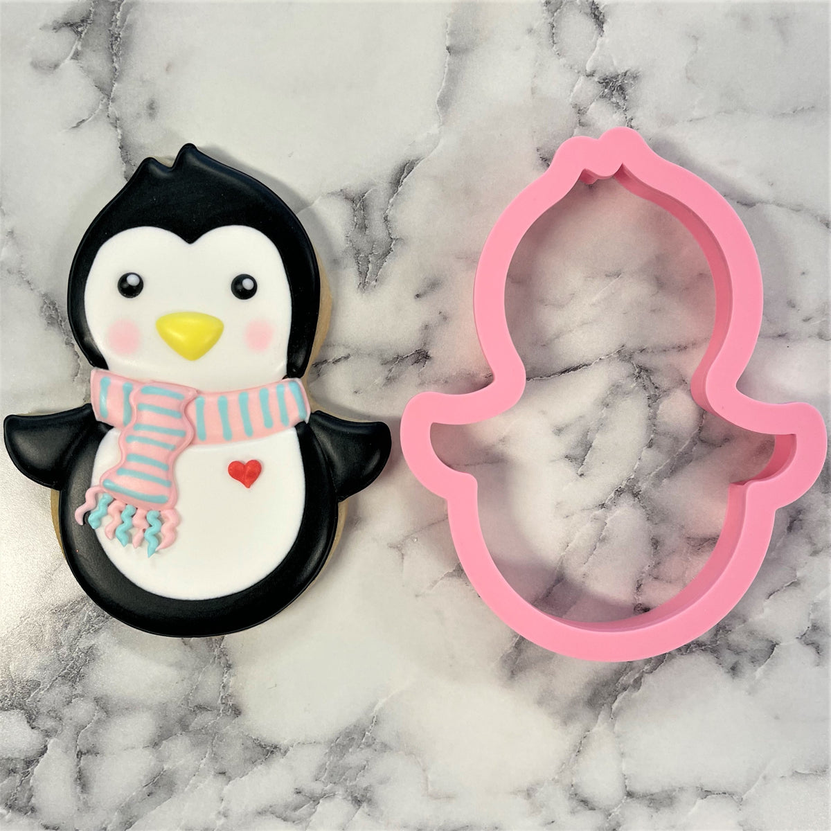 Pink silicone penguin roly poly cookie cutter next to decorated black and white penguin sugar cookie with scarf and red heart