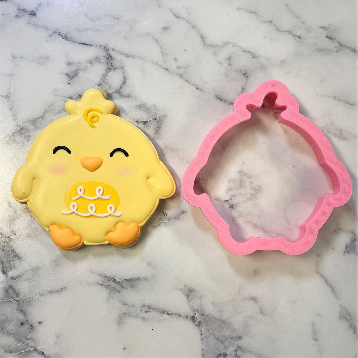 Yellow chick-shaped cookie with orange beak and feet pink cheeks white icing next to pink silicone round chick cookie cutter mold