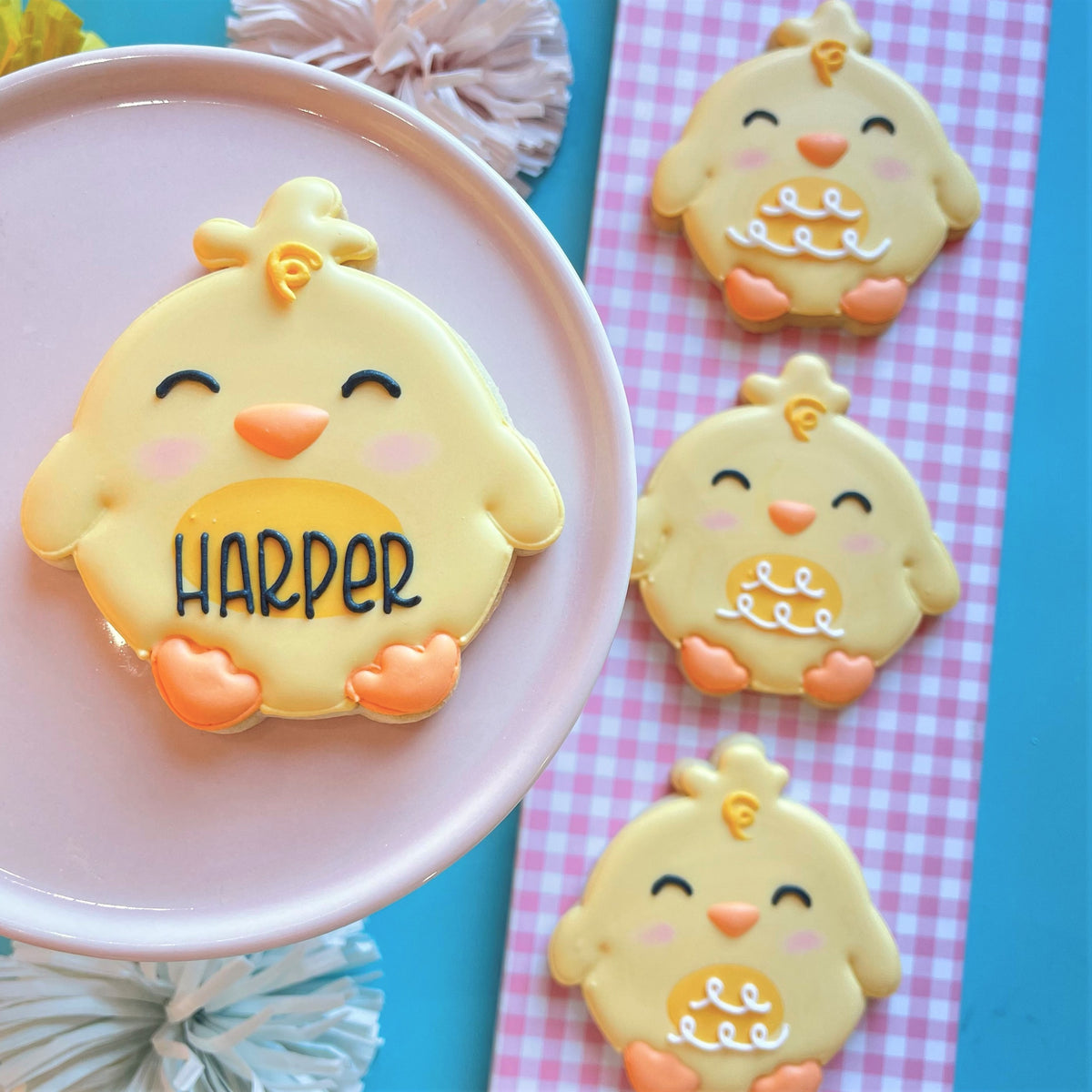 Yellow chick-shaped cookie with orange beak feet pink cheeks black icing eyes and harper on belly made using round chick cookie cutter