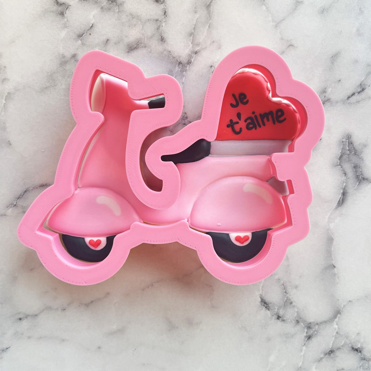 Back of a pink scooter-shaped metal cookie cutter with a red heart that reads je t’aime for baking use
