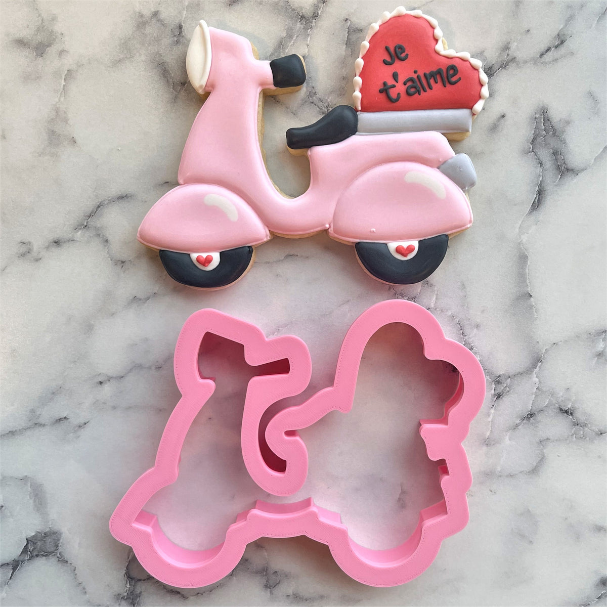 Pink scooter-shaped metal cookie cutter with matching pink sugar cookie decorated with icing and je taime on rear heart