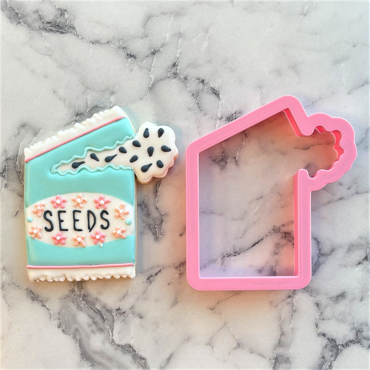 A pink plastic seed packet cookie cutter next to a decorated turquoise and white seed packet sugar cookie