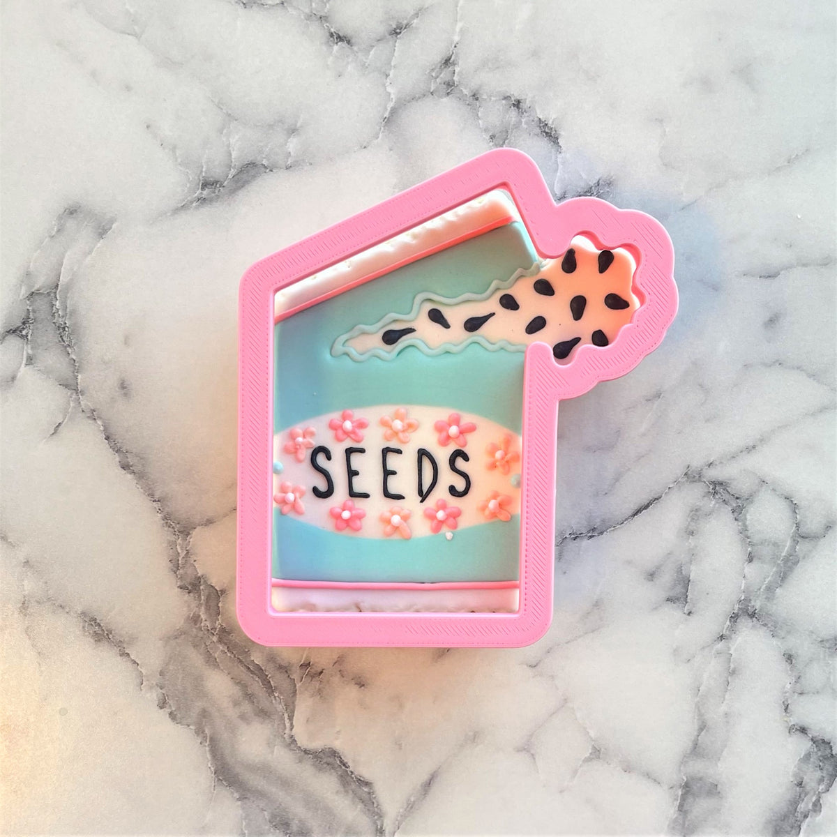 Pink plastic seed packet cookie cutter with teal interior black seeds lettering and pink floral decorations
