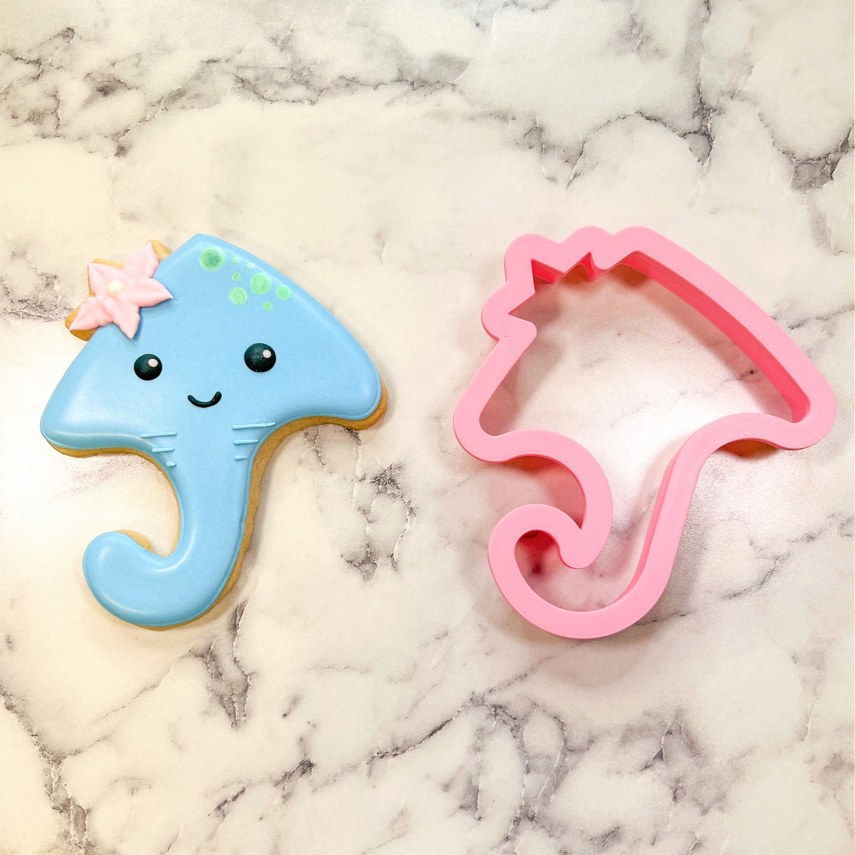 A light blue glossy cookie shaped like a smiling jellyfish with green polka dots and a pink starfish made with stingray flour box club cookie cutter