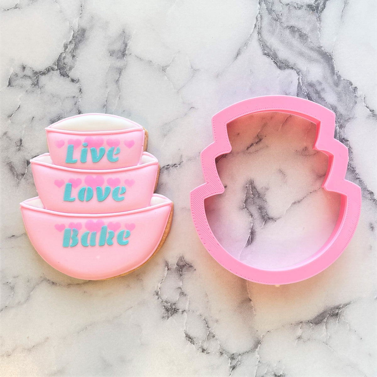 Pink silicone stacked bowls cookie cutter shaped like tiered cake with live love bake teal icing and pink heart accents