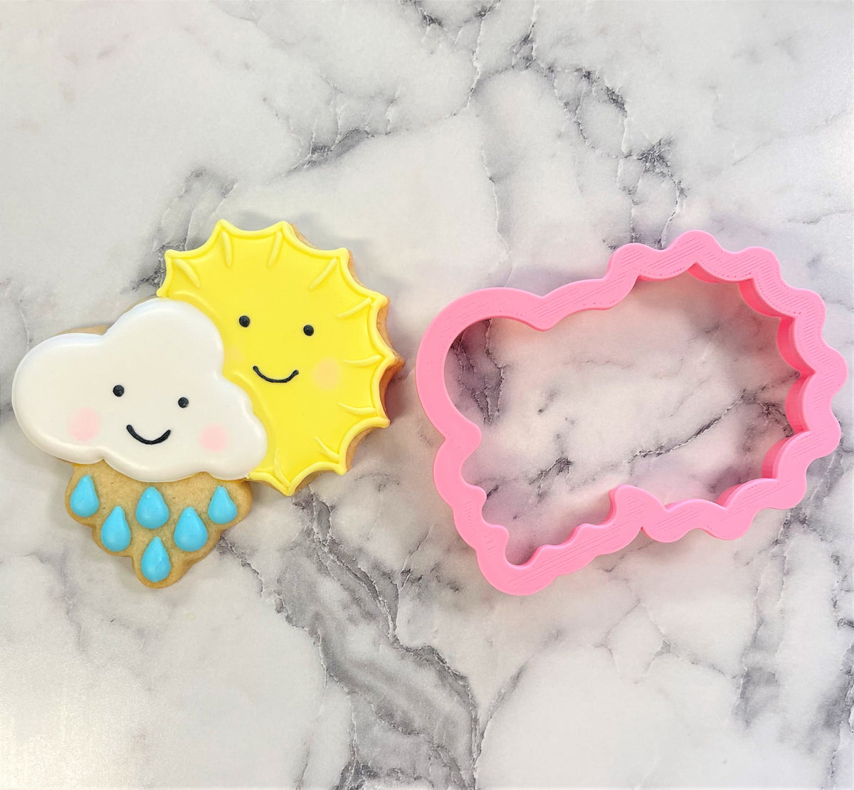 A pink plastic cloud-shaped cookie cutter with scalloped edge next to a decorated sun and cloud sugar cookie