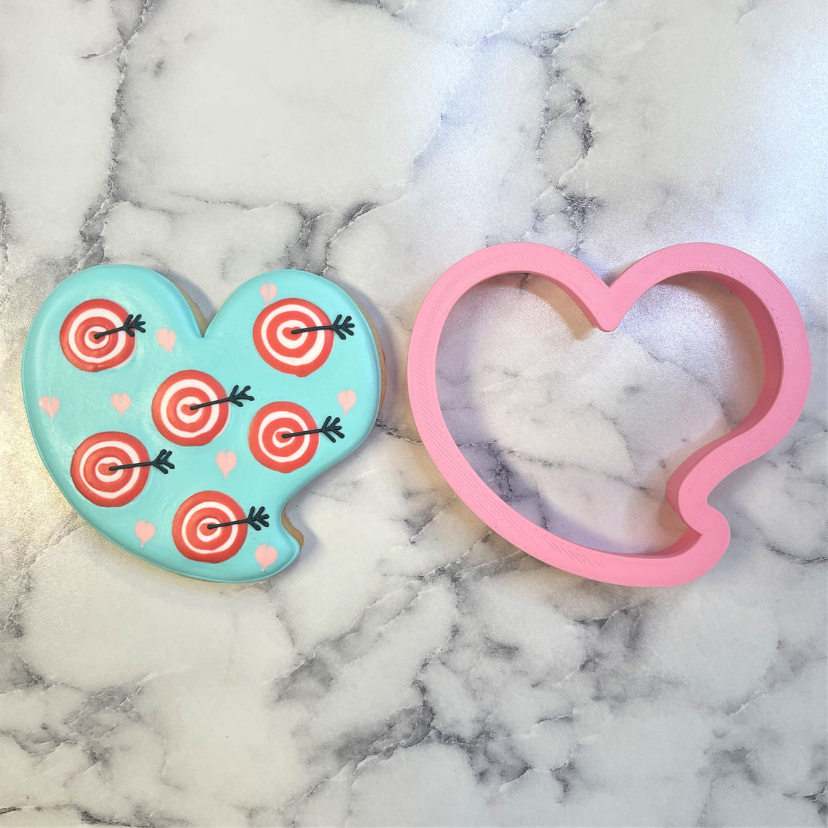 A pink heart talk bubble cookie cutter beside a light blue decorated cookie with red targets and black arrows