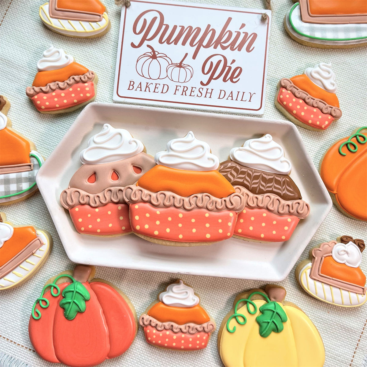 A white hexagonal plate with three pumpkin pie shaped cookies decorated with orange pink icing and whipped cream toppers