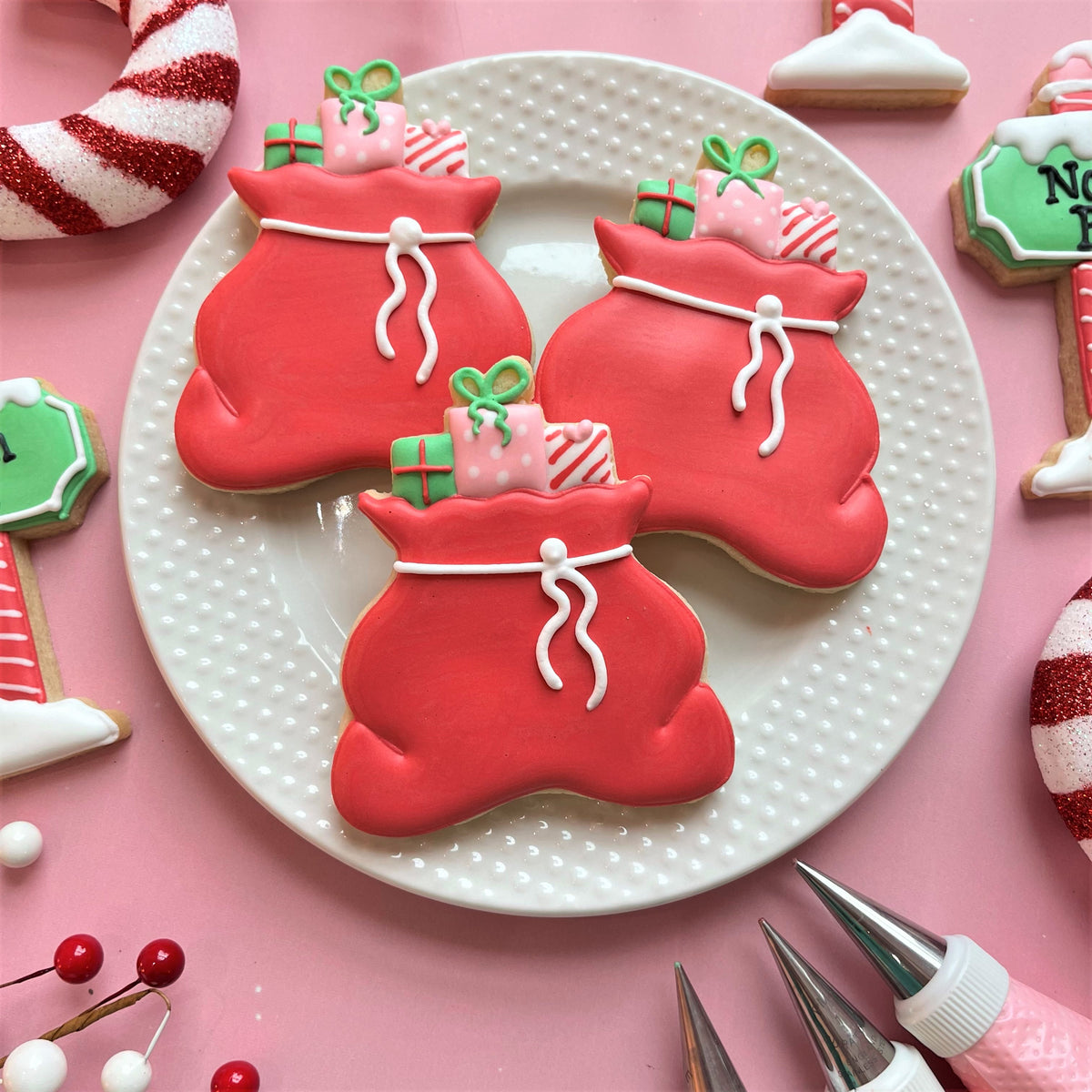 North pole cookie decorating class recording with red sugar cookies shaped like christmas gift sacks decorated with white icing ties and green pink gifts