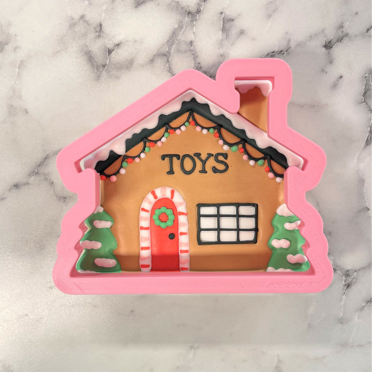 Pink toy workshop cookie cutter shaped like a gingerbread house with brown facade red door and festive holiday details