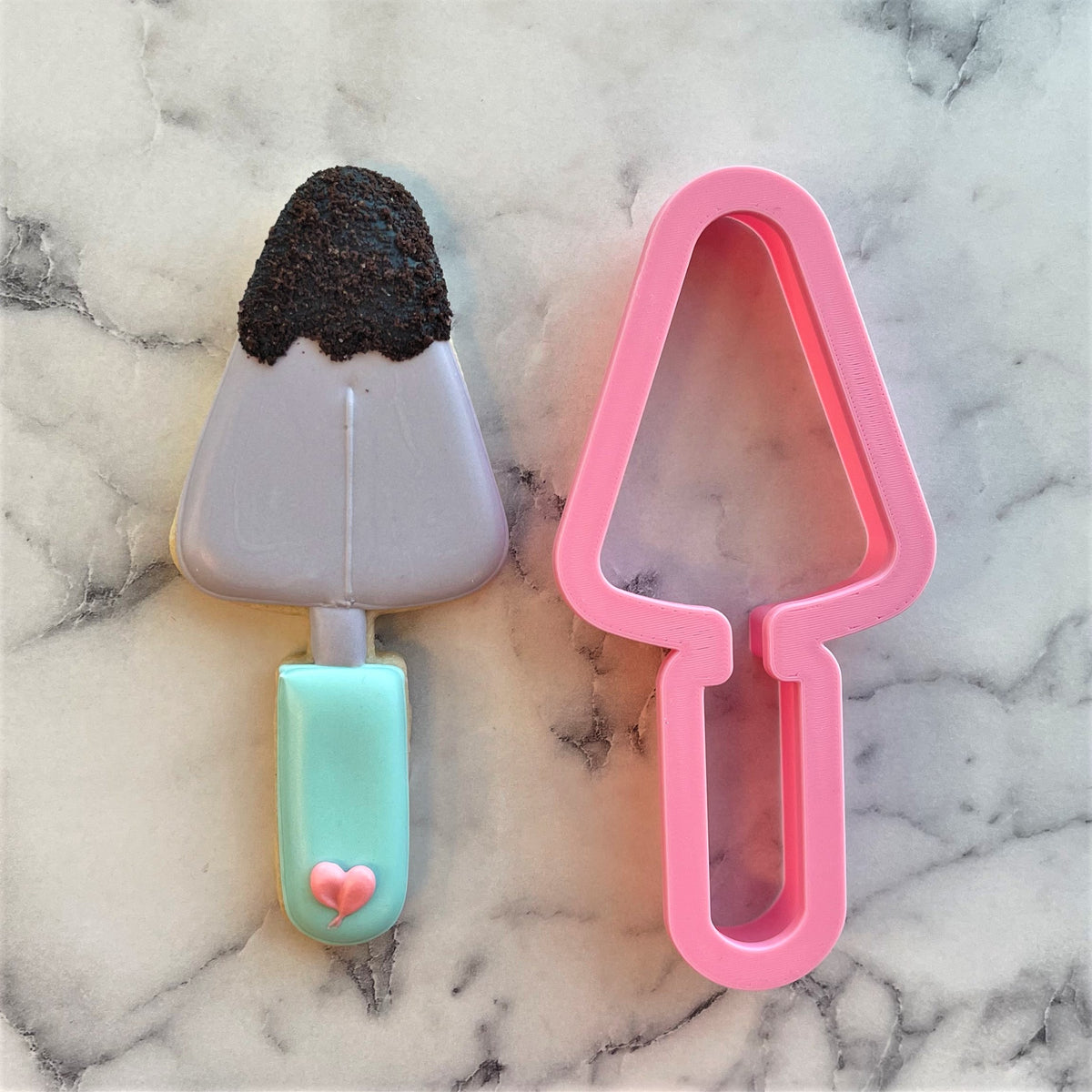 Pink plastic trowel cookie cutter next to chocolate popsicle cookie with mint green stick and heart detail