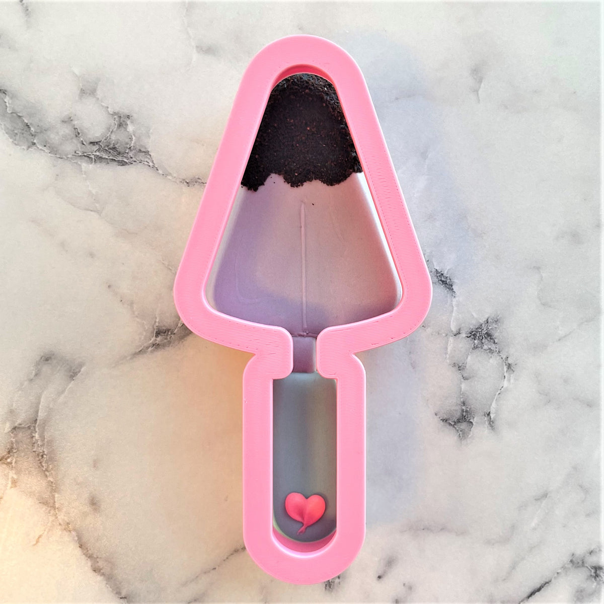 Pink plastic trowel cookie cutter with black chocolate-like filling and small pink heart detail at the bottom