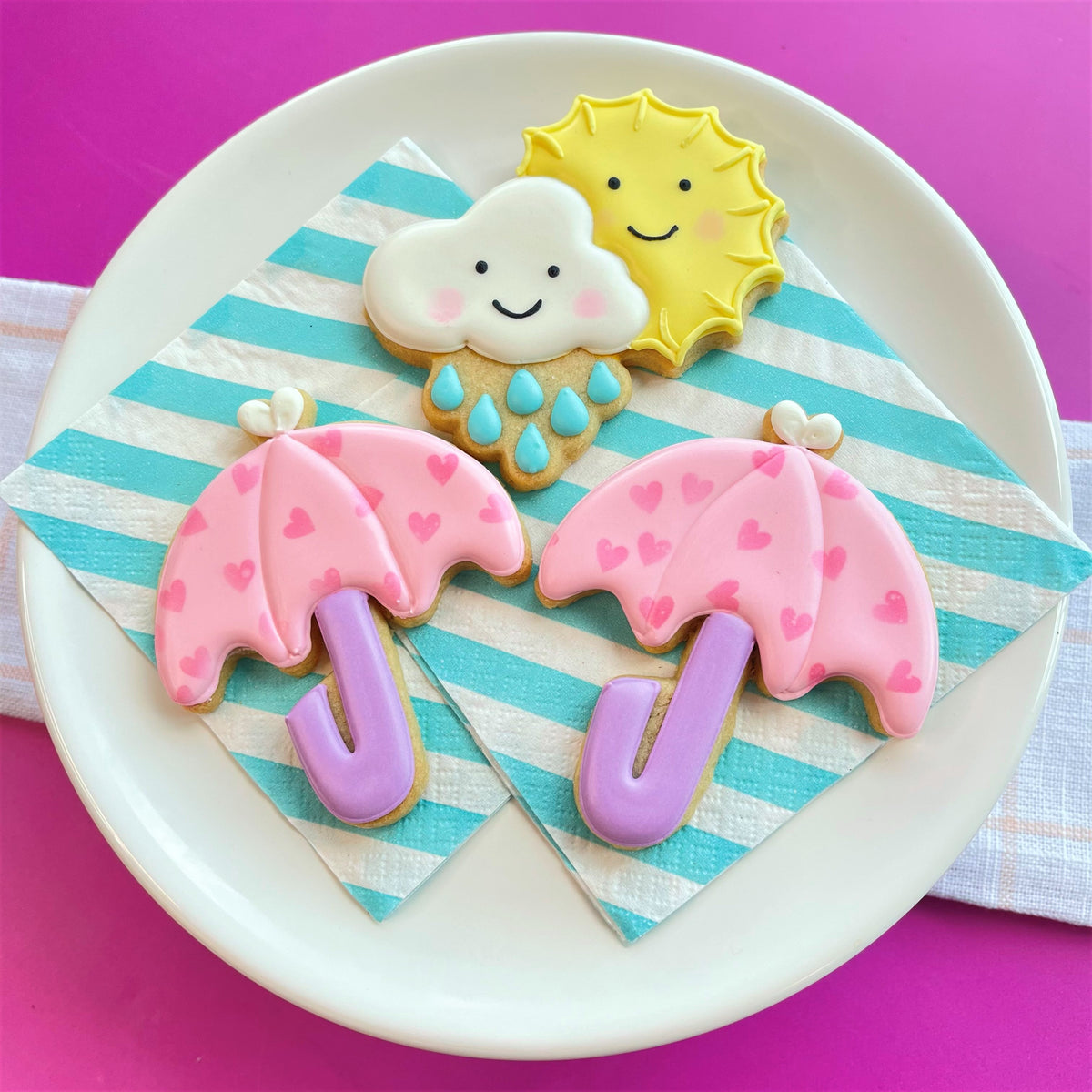 Pink heart-patterned umbrella cookies with purple handles plus smiling white cloud and yellow sun for spring cookie class