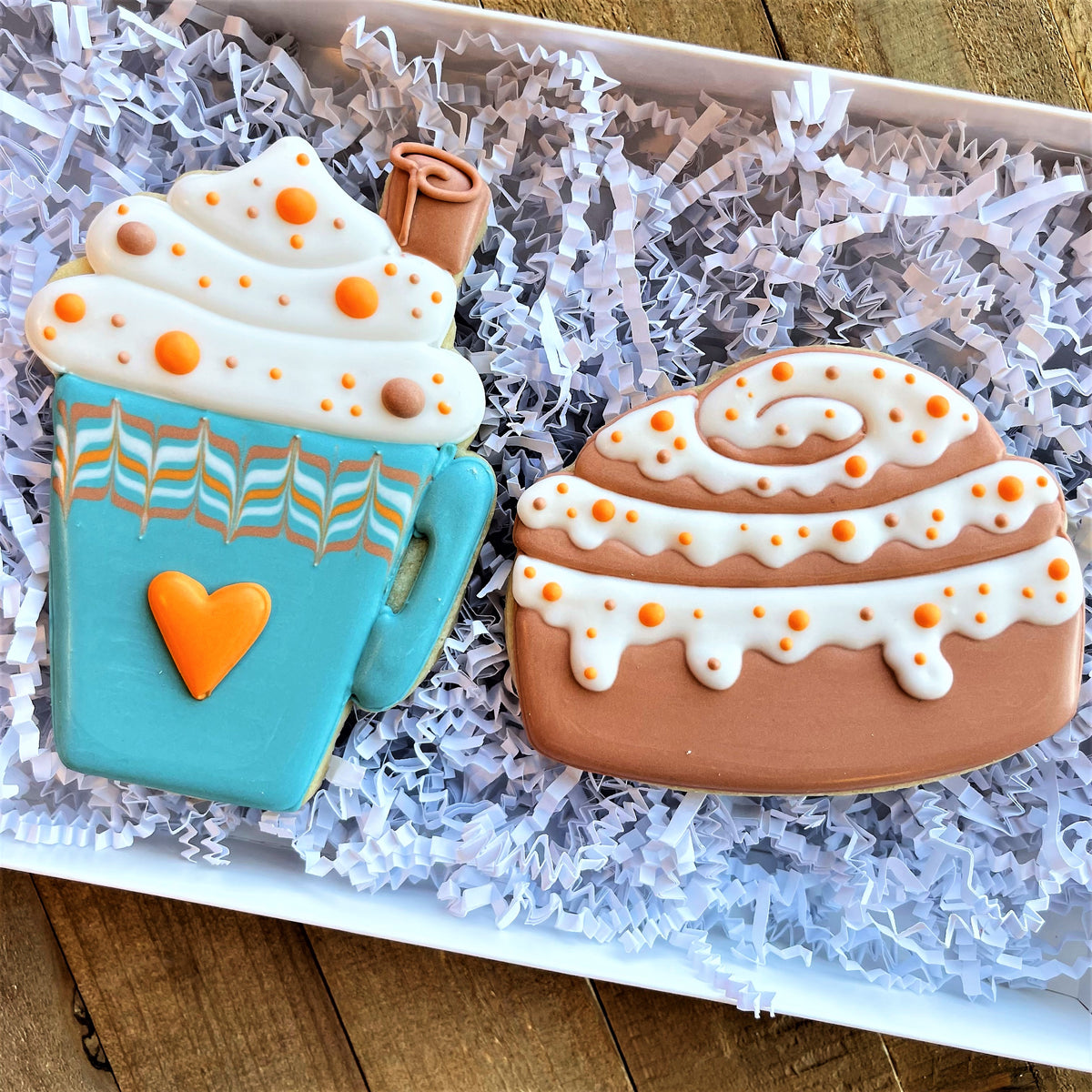 Two decorated sugar cookies shaped like a blue latte cup with whipped cream and a brown cinnamon roll with white icing drizzle