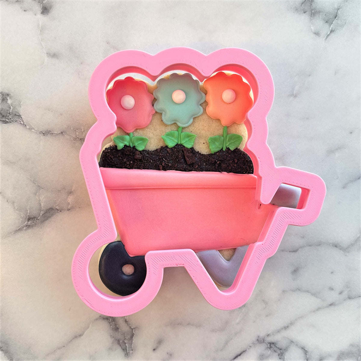 Pink plastic wheelbarrow cookie cutter filled with three colorful flower-shaped cookies for baking fun