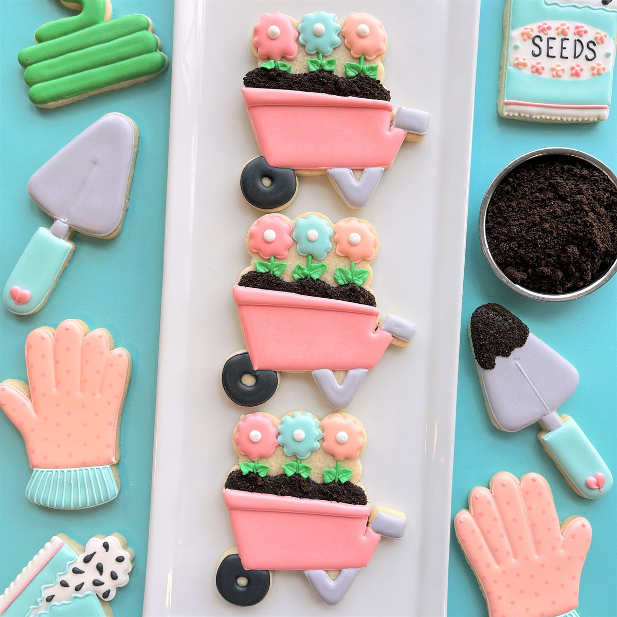 Pink sugar cookie wheelbarrows decorated with colorful flower cookies and dark brown edible dirt made with wheelbarrow cookie cutter