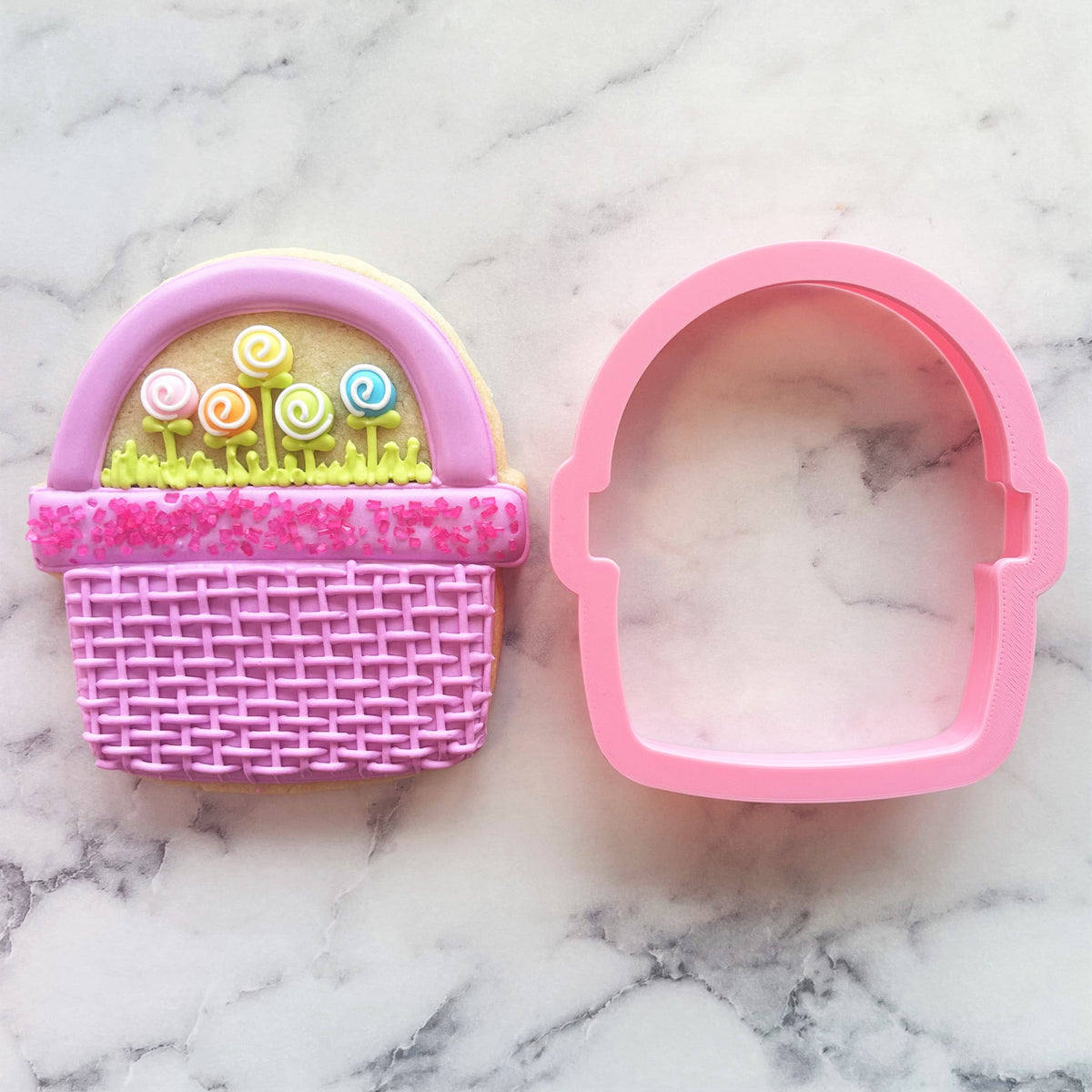 Wide basket cookie cutter in pink plastic with woven texture and handle beside a matching decorated cookie