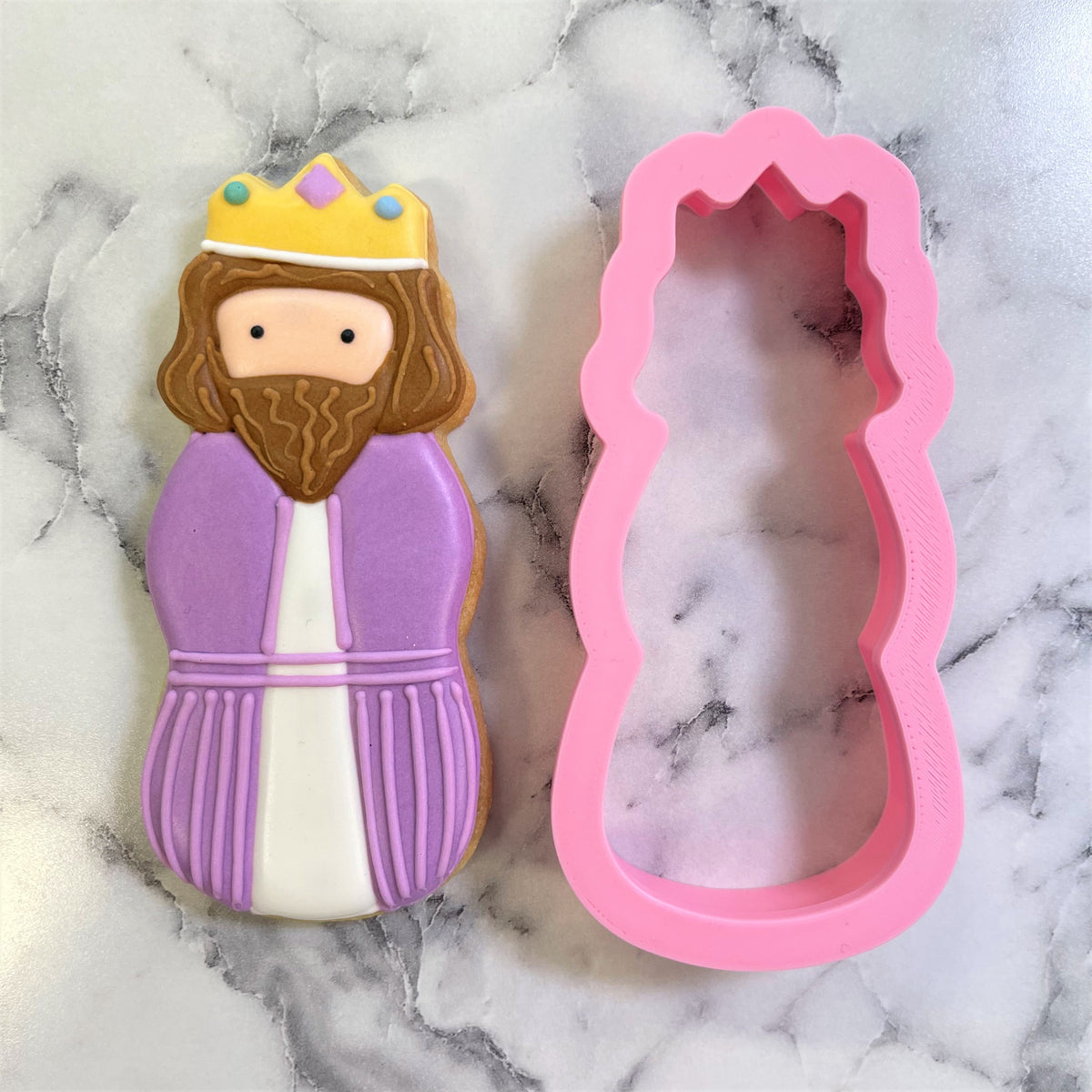 A purple and white frosted wiseman cookie with yellow crown beside a pink silicone cookie cutter mold