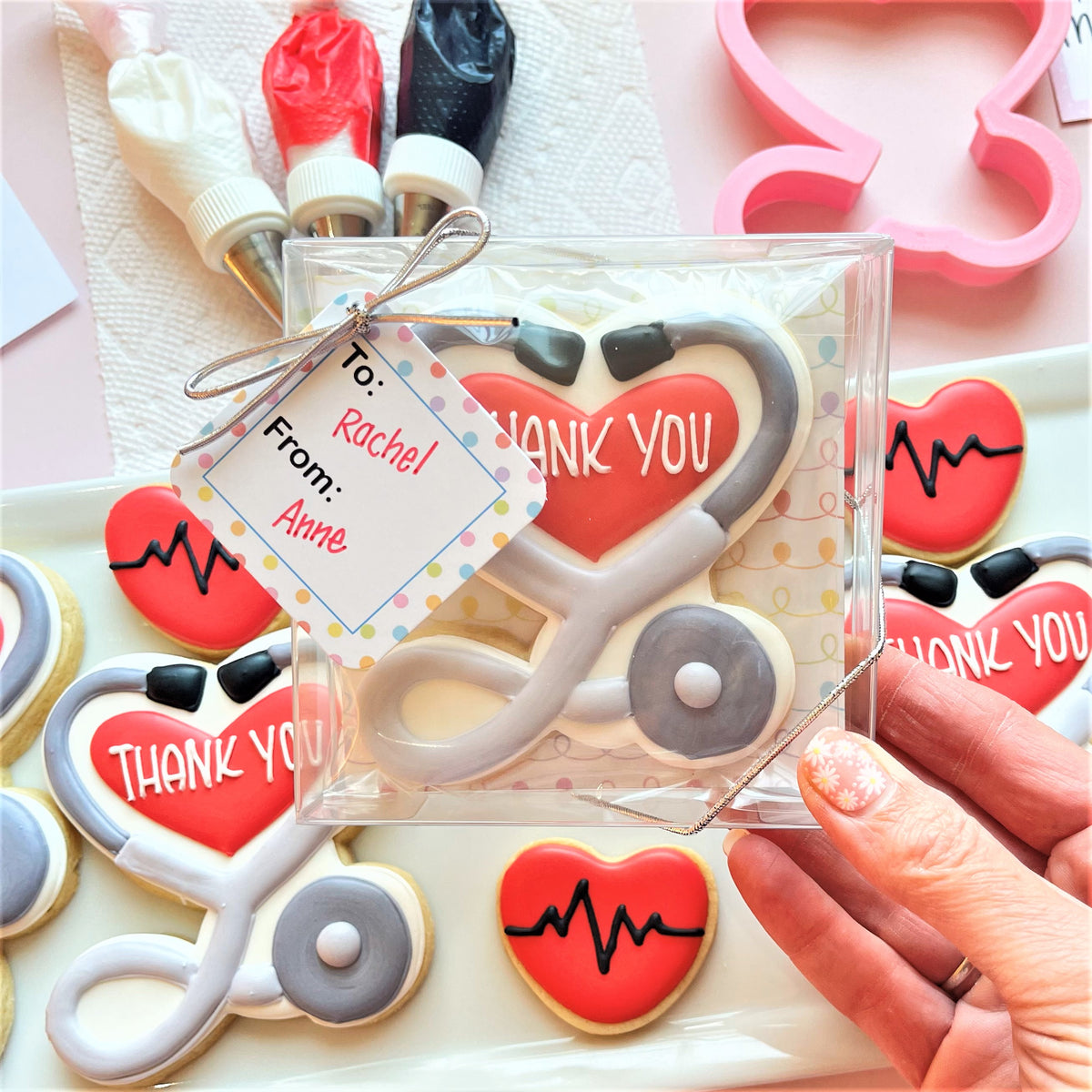 A red and gray stethoscope-shaped cookie with white thank you icing packaged in a clear plastic box for medical gift use