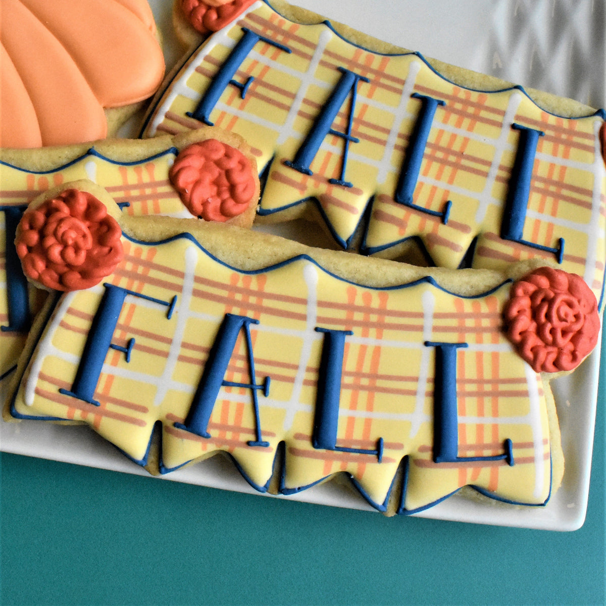 Yellow and orange plaid sugar cookies shaped like a banner with blue lettering spelling fall and red rose decorations for thanksgiving class recording