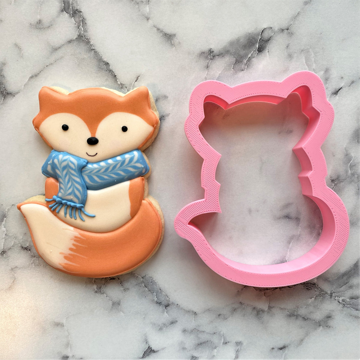 A pink fox-shaped metal cookie cutter next to a decorated sugar cookie with orange icing and a blue scarf