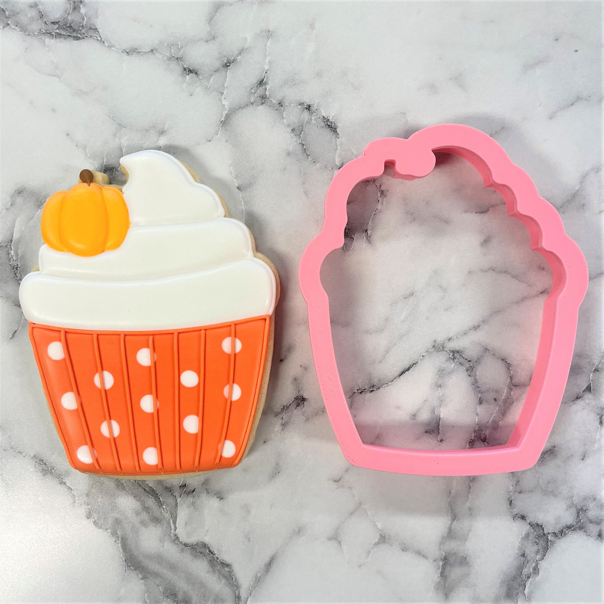 A pink silicone scalloped edge pumpkin cupcake cookie cutter beside an orange polka-dotted decorated sugar cookie