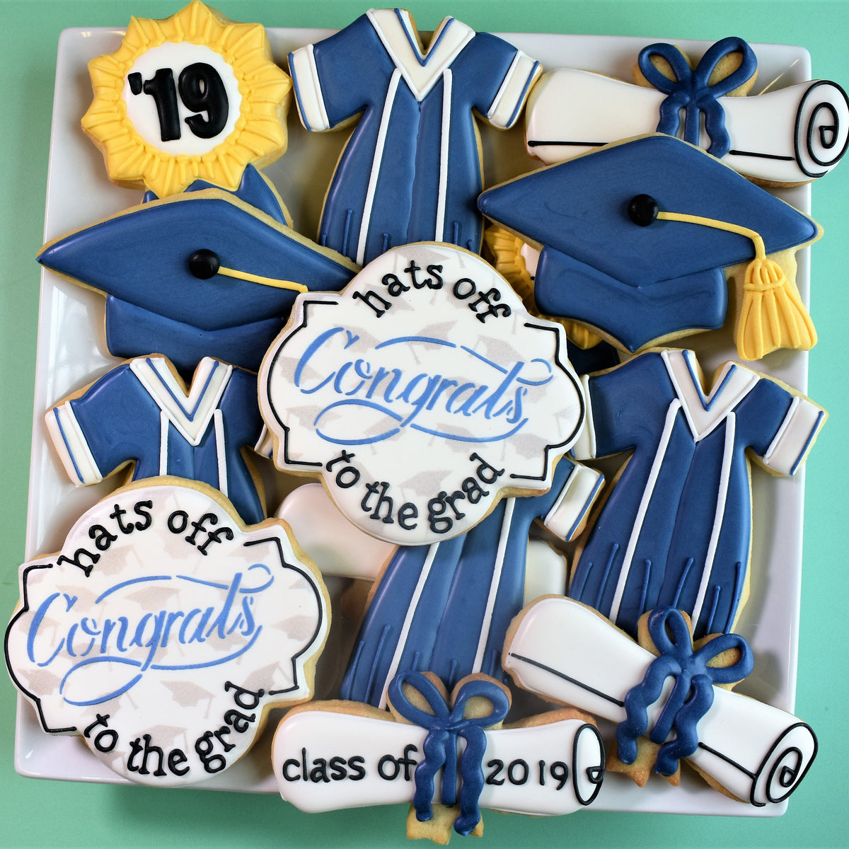 Blue and white graduation-themed sugar cookies shaped like gowns caps diplomas and a sun with 19 in grad cookie kit